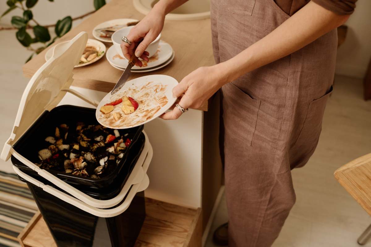 Low section of young modern woman with dirty plate putting food leftovers in bin with compost after eating to save environment